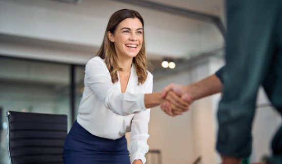 Happy mid aged business woman manager handshaking greeting client in office. Smiling female executive making successful deal with partner shaking hand at work standing at meeting table.