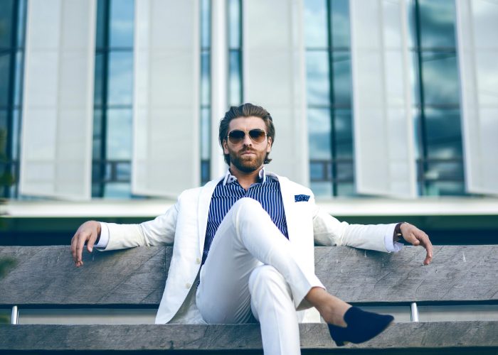 Fashionable man in white suit sitting confidently on a bench outdoors, embodying elegance and modern lifestyle.